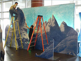 Workers on ladders assemble full size completed 3D mountain and castle display with lighting.